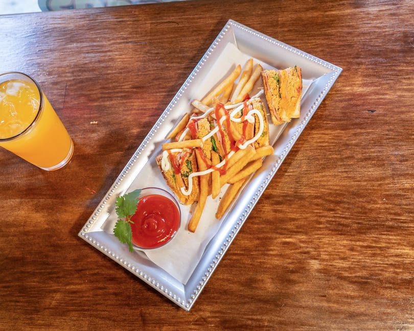A silver tray holds a club sandwich with fries, ketchup, and mayonnaise, next to a glass of orange juice.