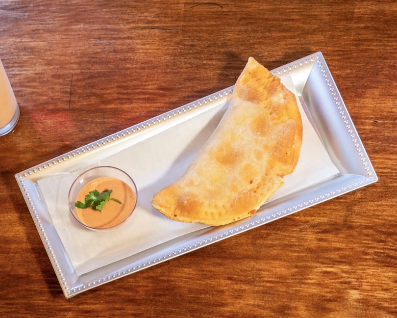 A golden-brown empanada with a side of dipping sauce garnished with cilantro on a silver tray.