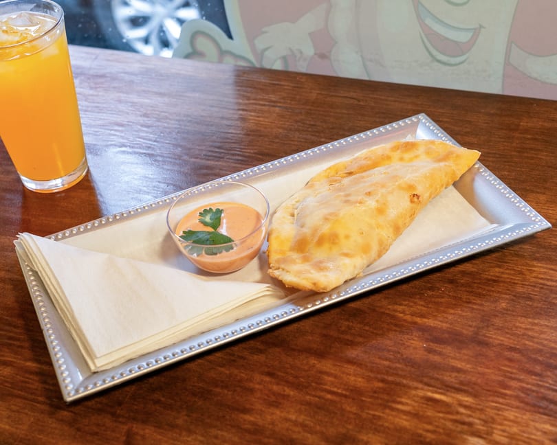Golden-brown empanada with dipping sauce and orange juice on a wooden table.