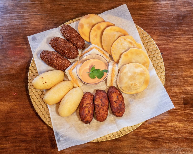 Assortment of fried appetizers including empanadas, croquettes, and corn fritters with a dipping sauce.
