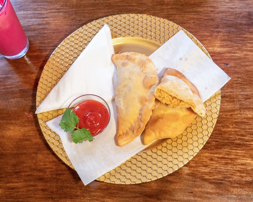 Three golden-brown empanadas on a gold honeycomb plate with ketchup and cilantro, next to a pink drink.