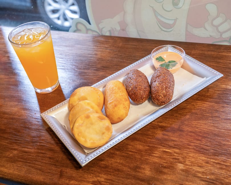 A silver tray holds fried pastries, croquettes, and a dipping sauce, with a glass of orange juice.