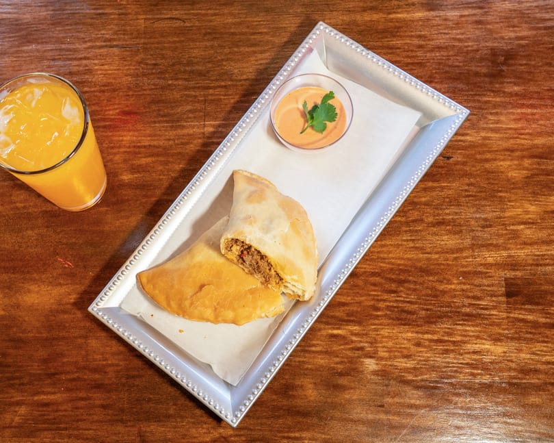 Two empanadas, one cut open to show the filling, with dipping sauce and orange juice.