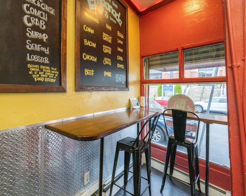 A small restaurant interior with a bar-height table, two stools, and menu boards on the wall.