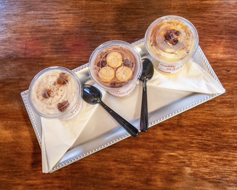 Three plastic cups of desserts with spoons on a silver tray, on a wooden table.