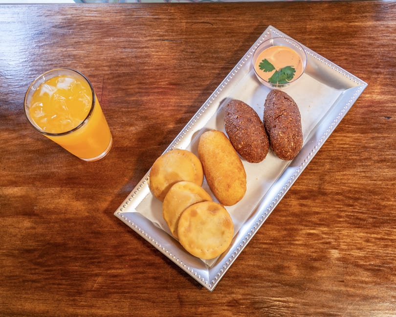 A silver tray with various fried pastries and a dipping sauce, next to a glass of orange juice.