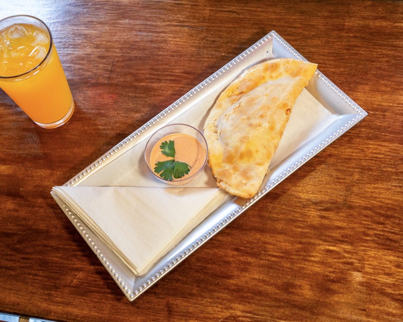 A golden-brown empanada with dipping sauce and an orange drink on a wooden table.