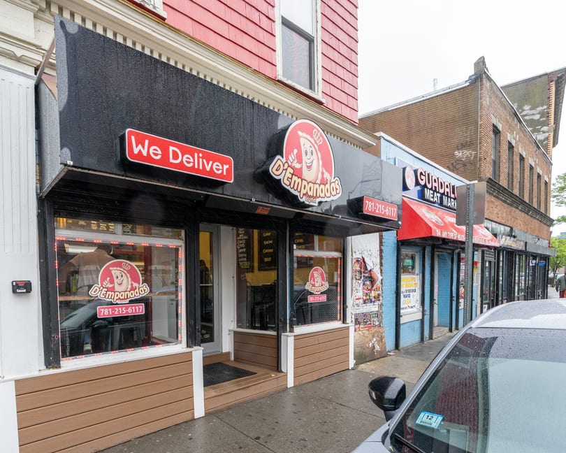 Exterior of D'Empanadas restaurant with a "We Deliver" sign and a car parked nearby.
